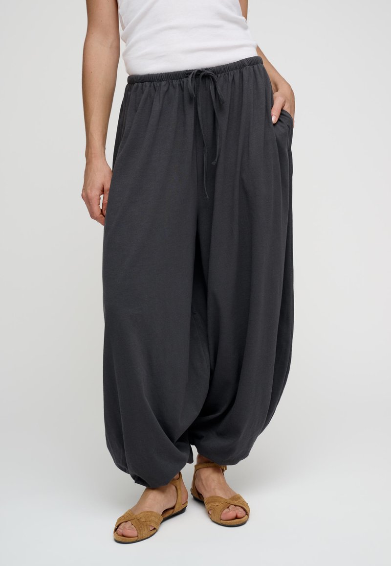 Pantalon harem noir à coupe décontractée avec taille élastique et cordon, porté avec des sandales ouvertes marron clair et un haut blanc sans manches.