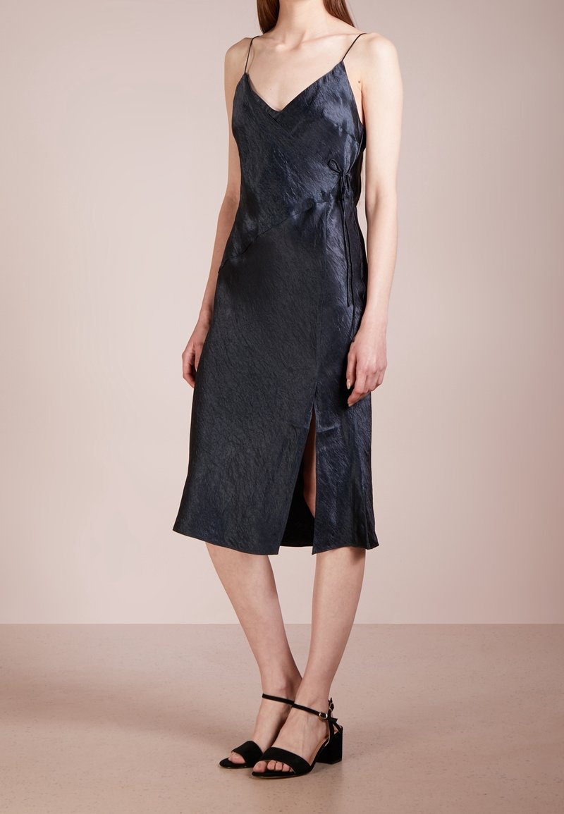 Robe midi en satin bleu marine avec un col en V, des bretelles spaghetti réglables, une fente latérale et un détail noué à la taille. Texture lisse.
