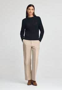 Un pull noir en maille torsadée avec des poignets et un ourlet côtelés, arborant un petit logo rouge. Assorti à un pantalon beigne taille haute et des bottines marron.