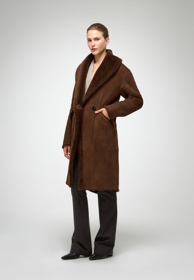 Pruun shearling-vatiga mantel, millel on lai, tekstuuriga krae, kaks eesmist taskut ja üks nupuga kinnituv sulgemine. Sobitatud tumevärviliste pükste ja kingadega.
