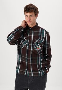 Brown plaid shirt with green and white accents, button-up front, two chest pockets, and a small branded patch on the left pocket.