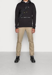 Czarny hoodie z logo, zestawiony z brązowymi spodniami i czarnymi botkami za kostkę. Strój uzupełnia czarna torba na ramię z żółtymi zamkami błyskawicznymi.