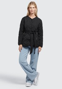 Giubbotto imbottito nero con cintura in vita annodata, dotato di scollo rotondo e chiusura con bottoni, abbinato a jeans a gamba larga blu chiaro e sneakers bianche.