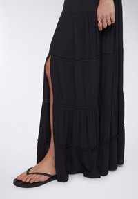 Abito maxi nero con design a strati, dotato di rifinitura in pizzo e uno spacco alto fino alla coscia. Abbinato a ciabatte nere, che mostrano le dita dei piedi e il piede.