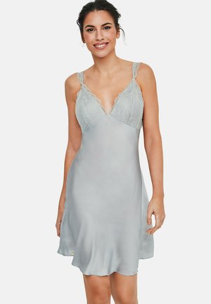 Femme portant une robe nuisette en satin gris clair avec bordure en dentelle et bretelles réglables, debout et souriante devant un fond uni.