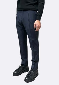 Pantaloni plissettati blu navy realizzati in materiale liscio. Abbinati a scarpe nere con lacci, superfici testurizzate e punte arrotondate.