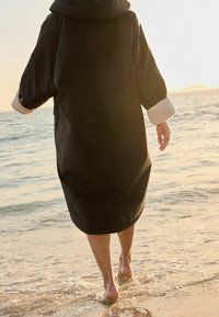 Zwarte waterdichte jas met een grote capuchon, voorzien van zachte witte manchetten, gedragen door een persoon die bij zonsondergang door ondiep water loopt.