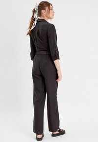 Tuta nera con design colletto, maniche lunghe arrotolate, vita cinturata e gambe larghe. Presenta un tessuto testurizzato e dettagli con bottoni.