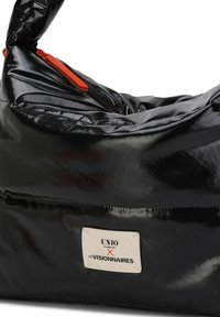 Bolsa acolchada negra y brillante con una cremallera naranja, que cuenta con una etiqueta prominente que dice "UNIO HAMBURG X LES VISIONNAIRES." Textura suave, forma redondeada.