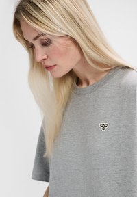 Grå bomullströja med rund hals och korta ärmar, med en liten svart och vit broderad logotyp på bröstet.