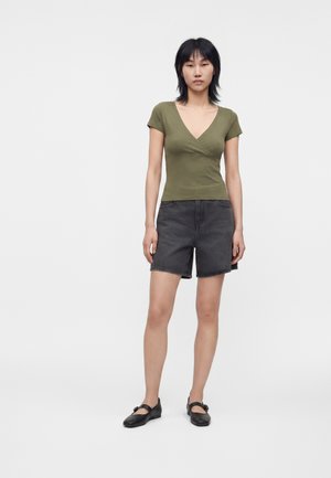 Femme debout face à l'avant portant un haut ajusté vert olive à col en V, un short en jean noir et des chaussures Mary Jane noires, sur un fond uni.