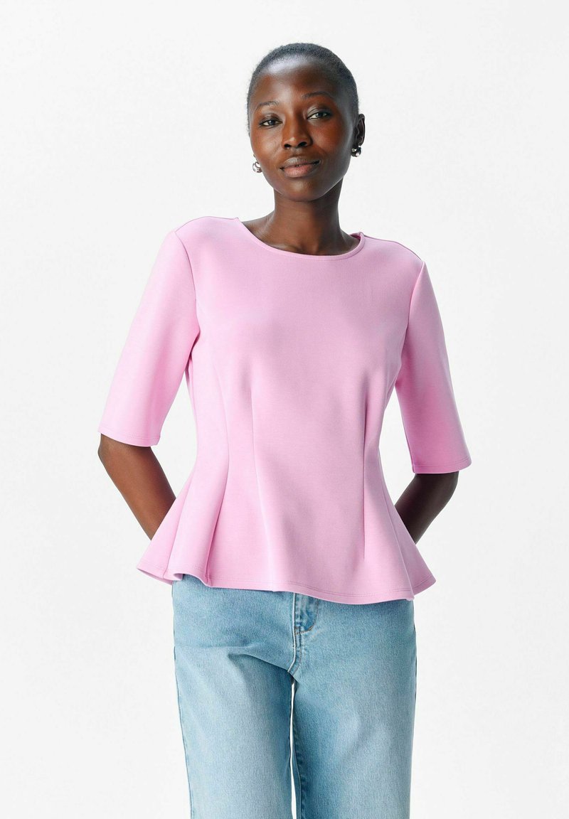 Rosa peplumtop med rund halsringning och trekvartslånga ärmar, tillverkad av ett mjukt material, i kombination med ljusblå denimjeans.
