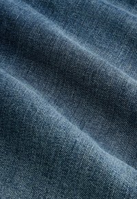 Tkanina dżinsowa w średnim odcieniu niebieskiego, charakteryzująca się delikatną fakturą z ukośnym splotem i miękkim układem, ukazująca naturalne zróżnicowania koloru.