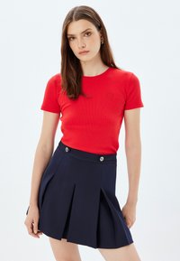 Camiseta roja ajustada de manga corta con textura acanalada y un logotipo sutil. Combinada con una falda plisada azul marino con detalles de botones plateados.