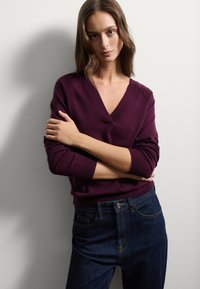 Eine Frau mit heller Haut und braunen Haaren trägt einen dunkelvioletten V-Ausschnitt-Pullover und dunkelblaue Jeans. Sie steht mit verschränkten Armen vor einer schlichten Wand.