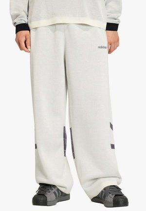 Pantaloni bianchi a gamba larga con logo Adidas nero sulla coscia, indossati con sneaker Adidas grigie e nere.