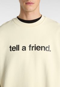 Sudadera color crema con cuello redondo y texto negro que dice "dile a un amigo". La textura parece suave y lisa.