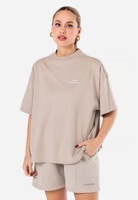 Beige oversized t-skjorte laget av myk bomull, med rund hals og korte ermer. Detaljen inkluderer subtil hvit tekst på brystet.
