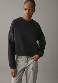 Svart sweatshirtt med fransdetaljer på ärmarna, rund halsringning och kort design, i kombination med ljusgrå jeans med sidostudsar.