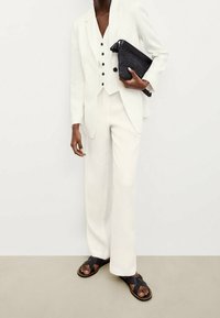 Henkilö, joka on pukeutunut täysin valkoisiin muotitakkisiin pukuun, mustiin sandaaleihin ja pitää mustaa teksturoitua clutch-laukkua kevyttä vaaleaa taustaa vasten.