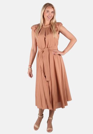 Lächelnde Frau mit langem blondem Haar trägt ein ärmelloses, tailliertes, hellbraunes Midikleid und beige Absatzsandalen mit Knöchelriemen, steht mit einer Hand in der Hüfte.