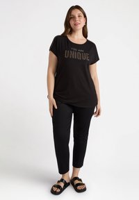 Camiseta negra con el eslogan "ERES ÚNICO" en purpurina, de mangas cortas, combinada con pantalones ajustados negros y sandalias negras simples.