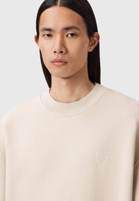 Felpa beige con collo a girocotola ribbed, caratterizzata da un discreto logo ricamato sul petto. Il tessuto ha una texture liscia.