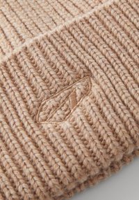 Knit beanie in a light beige shade, featuring a textured ribbed pattern and a small embroidered design resembling lips.