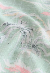 Tissu vert clair avec une texture tissée et un imprimé floral blanc, noir et pastel, présenté avec de doux plis créant des ombres douces.