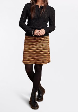 Kvinde iført sort strikket cardigan, mønstret orange og gul nederdel, sorte strømpebukser og sorte snørestøvler, stående med hænderne foldet.