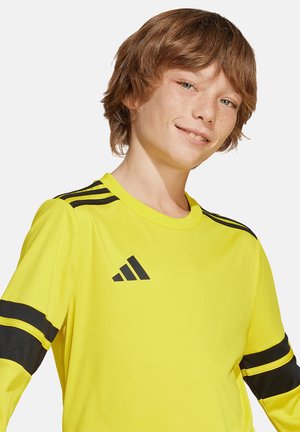 Giovane sorridente con lentiggini chiare che indossa una maglietta sportiva gialla brillante Adidas con strisce nere sulle maniche e sulle spalle.