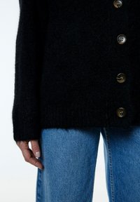 Schwarze Strickjacke mit großen braunen Knöpfen, einem gerippten Saum und langen Ärmeln, kombiniert mit hellblauen Jeans.