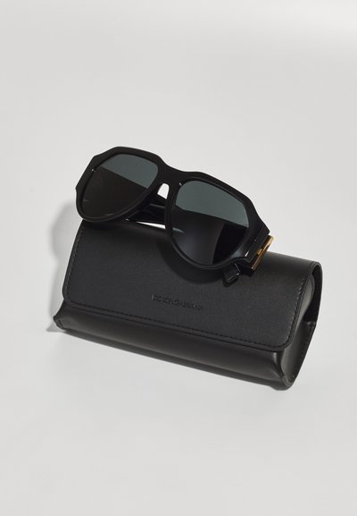Black sunglasses with angular frame and green lenses, resting on a black leather case with embossed logo. Smooth texture, sleek design.