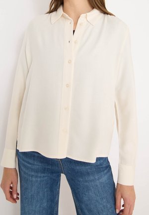 Person wearing a loose-fitting long sleeve cream button-up shirt and blue jeans, shown from neck to hips against a plain white background.