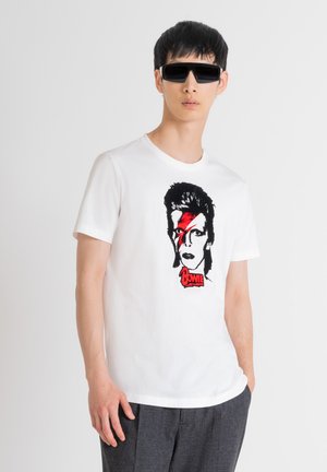 Camiseta blanca de algodón con un gráfico en negro y rojo de una cara con un rayo y el texto "Bowie" debajo. Modelo luciendo gafas de sol oscuras.