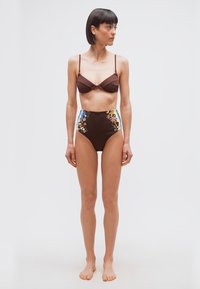 Γυναίκα με κοντά σκούρα μαλλιά, ντυμένη με κορυφή μπικίνι σε καφέ χρώμα και ψηλόμεσο καφέ κάτω μέρος μπικίνι με πολύχρωμα λουλουδάτα σχέδια, όρθια ξυπόλητη σε λευκό φόντο.