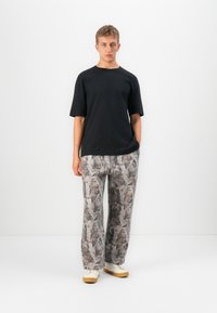 Tânăr în picioare, purtând un tricou negru supradimensionat, pantaloni largi gri cu un motive și adidași albi cu talpă de cauciuc.