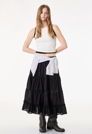 Młoda kobieta z długimi włosami, ubrana w biały, bezrękawnik, czarną spódnicę warstwową, paskowaną koszulę zawiązaną w pasie i czarne botki za kostkę.