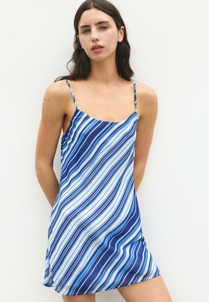 Femme aux cheveux foncés portant une robe sans manches rayée bleu et blanc sur un fond blanc uni.