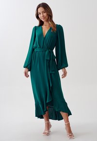 Zaļas apģērbs ar garām piedurknēm, jostu viduklim un pakāpeniski iegūtu apakšmalu. Auduma tekstūra ir nedaudz spīdīga, kas uzsver tā eleganto izskatu.
