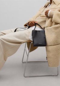 Svart läder axelväska med en slank rektangulär form, slät yta och korta remmar. Positionerad på en ljus stol mot en neutral bakgrund.