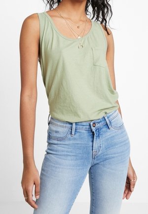 Kvinde iført lys grøn ærmeløs top med lille brystlomme og lyseblå højtaljede jeans, accessorized med lagdelte guldkæder.