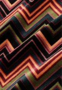 Tyger i flera färger i ett zigzag-mönster i nyanser av svart, grönt, orange och vinrött. Mjuk textur med synlig lugg och djup.