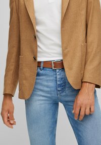 Bruine wollen blazer met twee voorzakken, zwarte knopen, gecombineerd met een wit shirt en blauwe spijkerbroek, wat een casual stijl tentoonstelt.