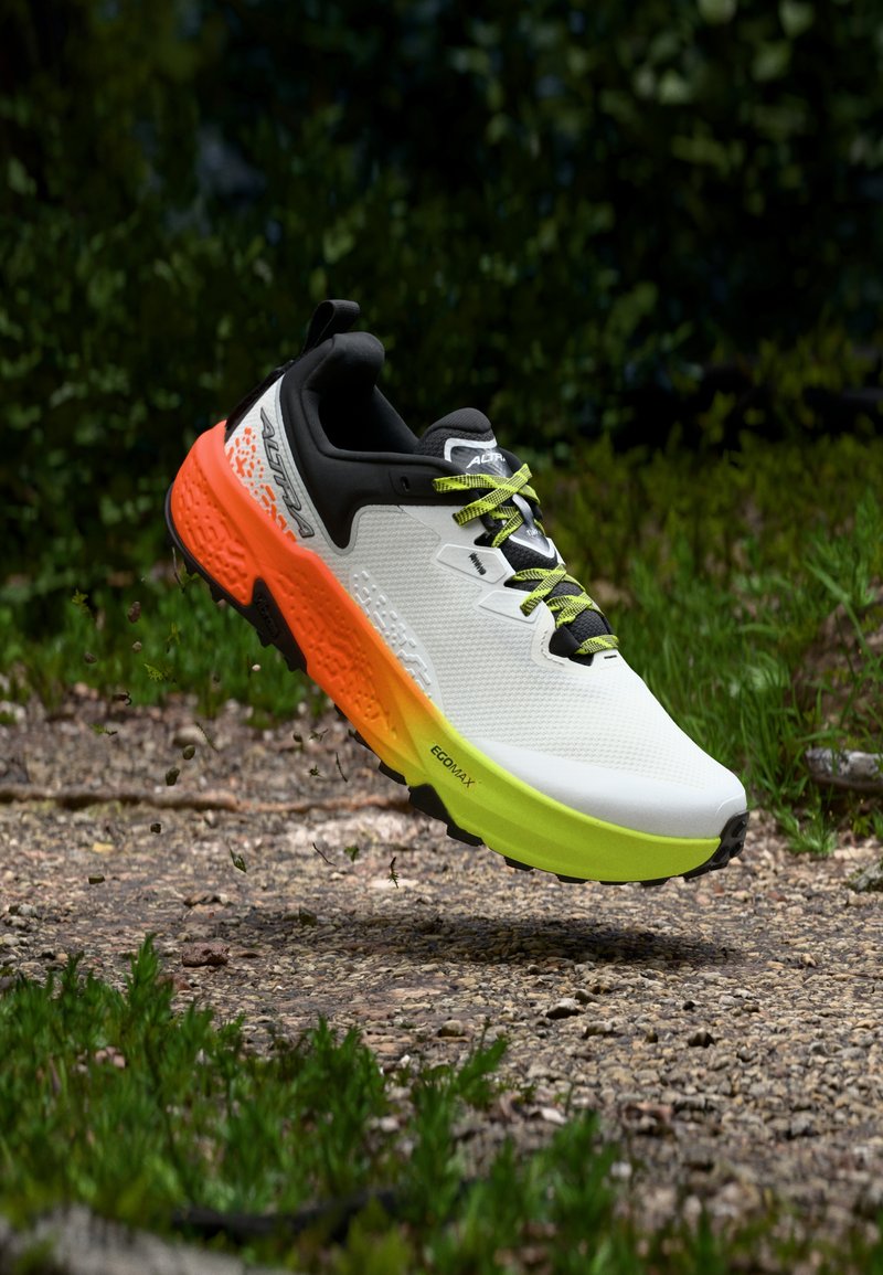 Chaussure de trail avec une tige blanche, un col noir, une semelle dégradée jaune fluo et orange, et des lacets verts, flottant au-dessus d'un chemin de gravier dans une forêt.