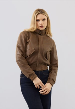 Frau trägt eine enganliegende braune Lederjacke mit hohem Kragen und elastischen Bündchen, kombiniert mit dunkelblauen Jeans, posiert vor einem einfarbigen Hintergrund.