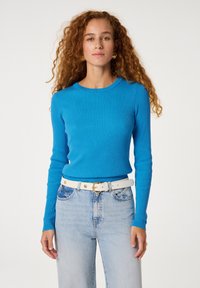 Blauwe geribde lange mouwen trui met een ronde halslijn, gecombineerd met lichte denim jeans en een witte riem met een goudkleurige gesp.