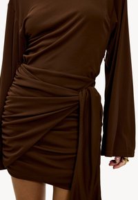 Robe marron à manches longues en matière douce, avec fronces à la taille et un détail noué, créant une silhouette ajustée.