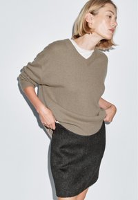 Maglione beige a collo a V con taglio morbido, abbinato a una mini gonna nera in feltro. Tessuto morbido con maniche arrotolate e indossato sopra una camicia bianca.