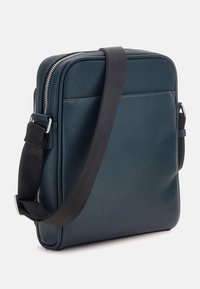 Mörk teal rektangulär väska med en texturerad yta, med en svart justerbar rem och silverfärgad dragkedja, slät yta med minimala detaljer.